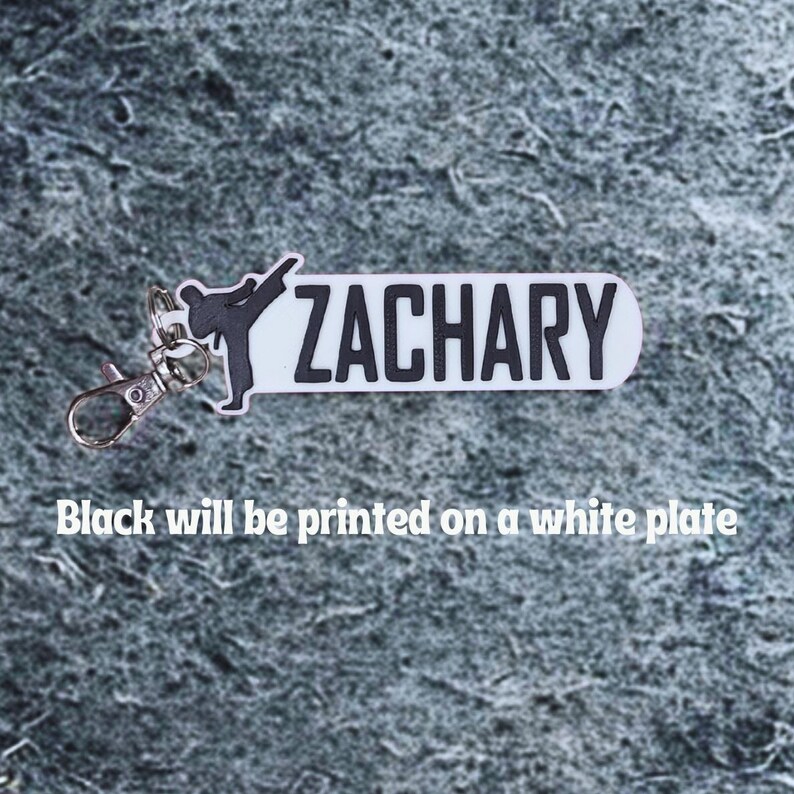 May include: A white keychain with the name "ZACHARY" in black block letters. A black silhouette of a person performing a martial arts kick is on the left. The text "Black will be printed on a white plate" is below.