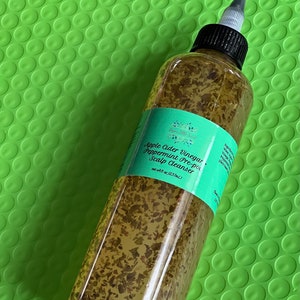 May include: A clear glass bottle with a black cap and a green label that reads "Apple Cider Vinegar Peppermint Pre-poo Scalp Cleanser" net wt 8 fl oz (237mL). The bottle is filled with a clear liquid and small green particles.