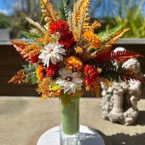 Puede incluir: Un arreglo floral con flores artificiales blancas, naranjas y rojo oscuro, mezcladas con tallos de trigo dorados y hojas de helecho verdes. Los tallos están insertados en un cilindro de espuma verde dentro de un florero de vidrio.