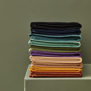 May include: A pile of folded corduroy fabric in a range of colours, such as black, navy, teal, green, purple, tan, orange, and rust. The fabrics are neatly stacked on a light green surface against a darker green background.