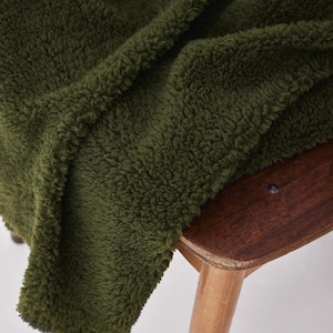 Può includere: Primo piano di una morbida coperta verde oliva con una superficie soffice e strutturata. La coperta è drappeggiata su una sedia di legno, evidenziando il suo aspetto accogliente. Il colore e la consistenza della coperta suggeriscono calore e comfort.