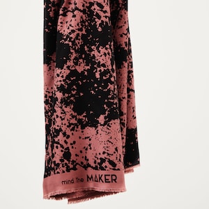 Può includere: Un tessuto rosa e nero con un design a spruzzo di vernice. Il tessuto ha un bordo a frangia e le parole "mind the MAKER" stampate in basso.