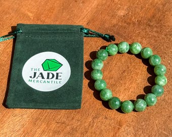 May include: A green jade bead bracelet next to a dark green drawstring pouch with a white circular logo that reads "THE JADE MERCANTILE". The bracelet is made of round, polished jade beads.
