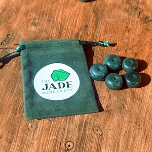 Könnte beinhalten: Grüne Jade-Donut-Perlen neben einem grünen Beutel mit Kordelzug. Der Beutel hat ein weißes rundes Etikett mit einem grünen geometrischen Design und dem Text "THE JADE MERCANTILE". Die Perlen liegen auf einer Holzoberfläche.