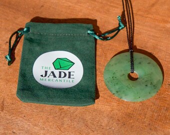 May include: A green jade pendant in the shape of a donut, hanging from a black cord. The pendant is on a wooden surface and comes with a green velvet pouch with the logo "The Jade Mercantile" printed on it.