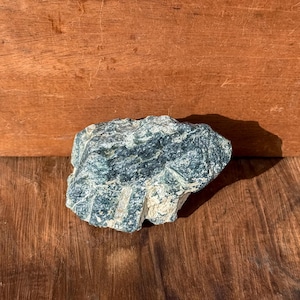 Op de afbeelding: Een ruwe, natuurlijke steen met een mix van donkerblauwgroen en gebroken wit. De steen heeft een textuur en onregelmatige vorm, rustend op een houten oppervlak. De kleurvariaties van de steen creëren een marmer effect.