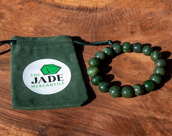 May include: A green jade bracelet with a dark green pouch that says "The Jade Mercantile" on a wooden surface.