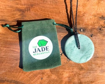 May include: A green jade pendant necklace with a round disc shape. The necklace is on a black cord and comes with a green velvet pouch with the logo "The Jade Mercantile".