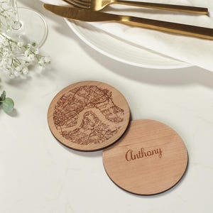 May include: Two circular wooden coasters. One has a detailed map design, the other is engraved with the name "Anthony". The coasters are light brown and rest on a white surface. The map appears to be of a city.