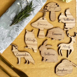 Op de afbeelding: Houten naambordjes in verschillende dierfiguren, waaronder een vos, een uil en een konijn, elk met een gegraveerde naam. De kaarten liggen op een gele ondergrond met een takje rozemarijn en een servet met patroon.