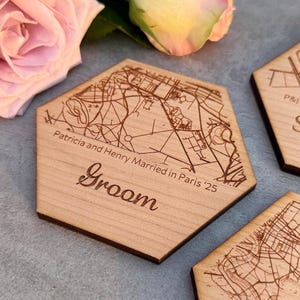 Op de afbeelding: Houten zeshoekige onderzetter met een kaartontwerp en het woord "Groom" gegraveerd. De onderzetter bevat ook de tekst "Patricia and Henry Married in Paris '25". De onderzetter is lichtbruin van kleur.