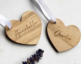May include: Two wooden heart-shaped ornaments with white ribbon hangers. One ornament has the name "Annabella" engraved, and the other has "Charlie." The wood grain is visible. A sprig of lavender is in the lower portion of the image.