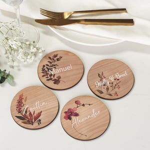 May include: Four round wooden coasters with personalized names and floral designs. Each coaster features a different name in white script: Manuel, Molly & Brad, Caitlin, and Alexander. Gold cutlery and a white plate are in the background.