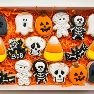 May include: A box filled with Halloween-themed sugar cookies. The cookies are decorated with various designs, including ghosts, pumpkins, candy corn, skeletons, and bats. The box is lined with orange shredded paper.