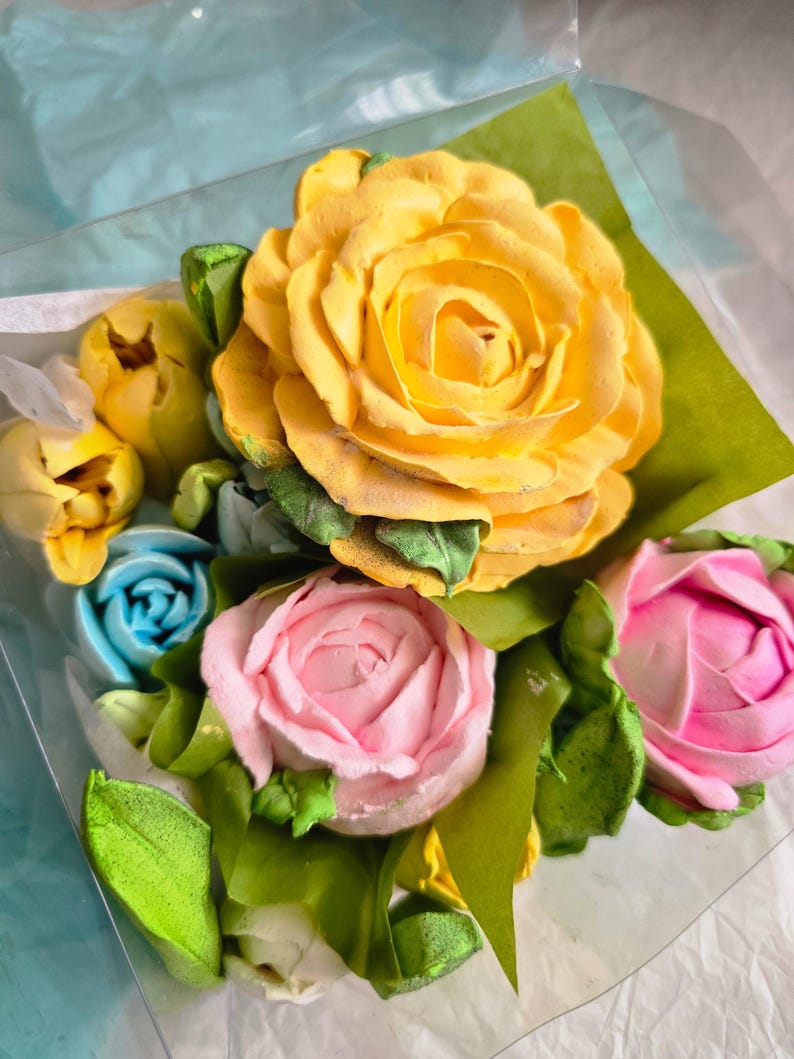 May include: A bouquet of colorful, edible flowers. The arrangement features a large yellow rose, pink and blue roses, and yellow buds, with green leaves. The flowers are presented in a clear plastic container.