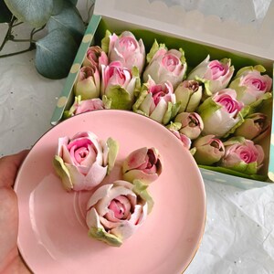 May include: A pink plate with three rose-shaped desserts, each with pink and white petals and green leaves. A box filled with similar desserts is in the background. The box has a clear window and a green and white polka dot design.