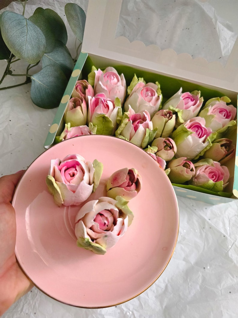 May include: A pink plate with three rose-shaped desserts, each with pink and white petals and green leaves. A box filled with similar desserts is in the background. The box has a clear window and a green and white polka dot design.