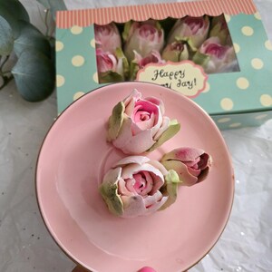 May include: A pink ceramic saucer holds three delicate, light pink and green rose buds. A box with a polka dot pattern and the words "Happy Day!" contains more roses. The roses have a soft, textured appearance, suggesting they may be made of paper or fabric.