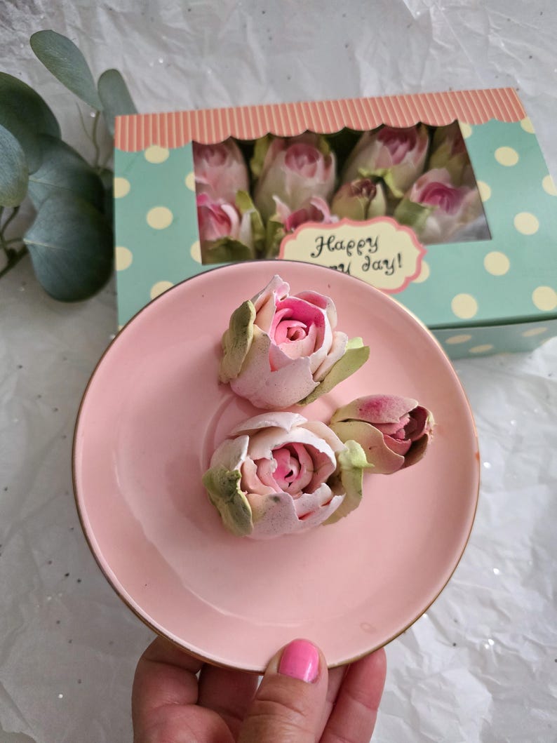 May include: A pink ceramic saucer holds three delicate, light pink and green rose buds. A box with a polka dot pattern and the words "Happy Day!" contains more roses. The roses have a soft, textured appearance, suggesting they may be made of paper or fabric.