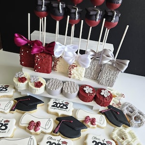 Op de afbeelding: Een assortiment traktaties met een afstudeerthema, waaronder cakepops met afstudeerhoeden, versierde koekjes met diploma's en afstudeerhoeden. De koekjes tonen de tekst "Class of 2025 congratulations!" in het zwart.