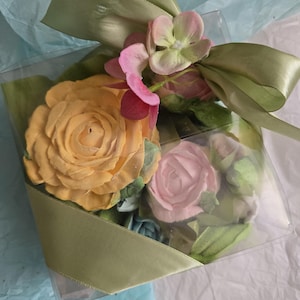 May include: A clear square box tied with a light green ribbon, filled with artificial flowers. The flowers include a large yellow rose, a pink rose, and smaller pink and green blossoms. The box sits on a bed of blue and white tissue paper.