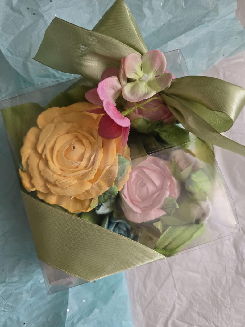 May include: A clear square box tied with a light green ribbon, filled with artificial flowers. The flowers include a large yellow rose, a pink rose, and smaller pink and green blossoms. The box sits on a bed of blue and white tissue paper.