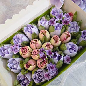 May include: A floral arrangement of pastel-colored flowers, including tulips and roses, presented in a white box with a clear lid. The flowers are in shades of purple, pink, and yellow, with green leaves. The arrangement is set against a white background.