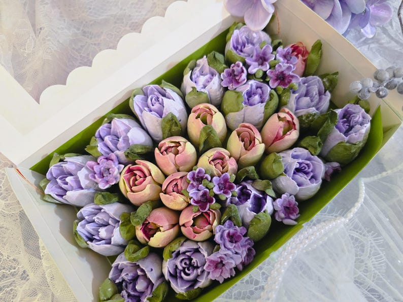 May include: A floral arrangement of pastel-colored flowers, including tulips and roses, presented in a white box with a clear lid. The flowers are in shades of purple, pink, and yellow, with green leaves. The arrangement is set against a white background.