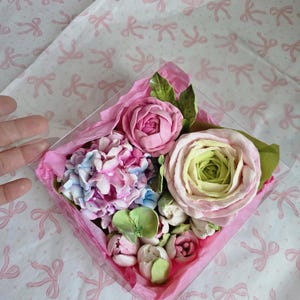 May include: A clear, square box filled with a variety of pastel-colored, handcrafted flowers. The arrangement includes pink roses, blue and pink hydrangeas, and green leaves, all nestled on pink tissue paper. The background is white with pink bow patterns.