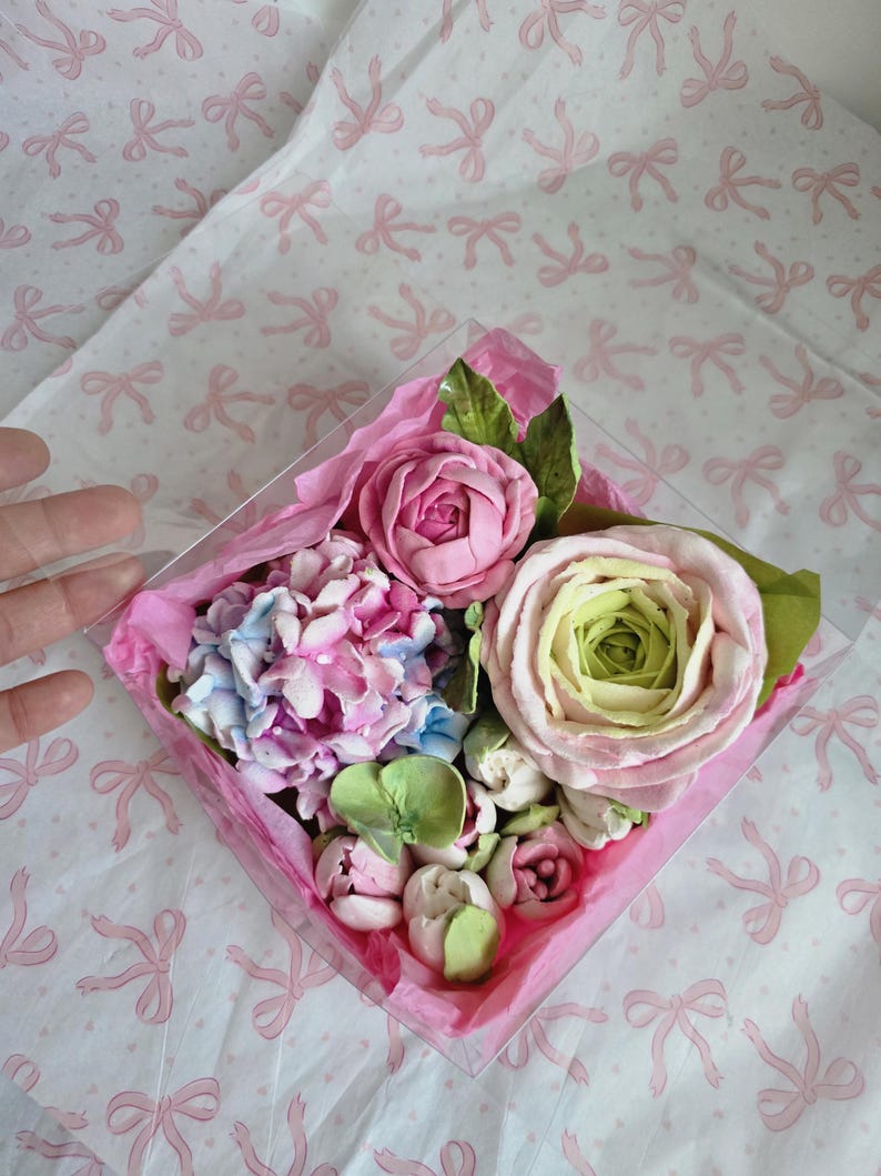 May include: A clear, square box filled with a variety of pastel-colored, handcrafted flowers. The arrangement includes pink roses, blue and pink hydrangeas, and green leaves, all nestled on pink tissue paper. The background is white with pink bow patterns.