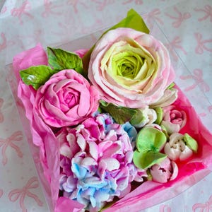 May include: A clear box filled with a colorful assortment of sugar flowers. The arrangement includes pink roses, a blue and pink hydrangea, and smaller white and pink buds, all nestled on pink tissue paper.