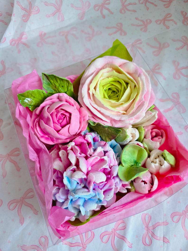 May include: A clear box filled with a colorful assortment of sugar flowers. The arrangement includes pink roses, a blue and pink hydrangea, and smaller white and pink buds, all nestled on pink tissue paper.