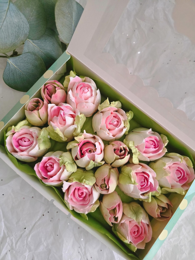 May include: A box of pink and white rose-shaped treats with green leaves. The treats are arranged in a green and white polka-dot box with a clear lid. The roses have a detailed design and are set on a bed of green paper.