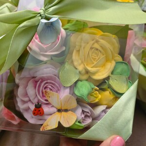 May include: A clear box filled with pastel-colored flower-shaped decorations, including roses and buds, accented with a ladybug and butterfly. The box is tied with a light green ribbon and bow.
