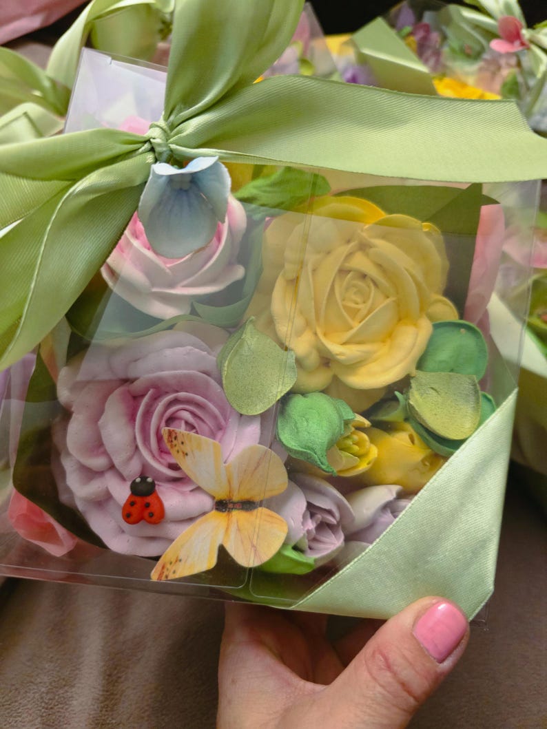 May include: A clear box filled with pastel-colored flower-shaped decorations, including roses and buds, accented with a ladybug and butterfly. The box is tied with a light green ribbon and bow.