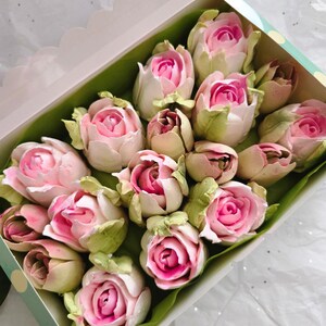 May include: A box of rose-shaped treats. The treats are pink and white, with green leaves. The box is white with a light green polka dot border. The treats are arranged on a green leaf-like paper.