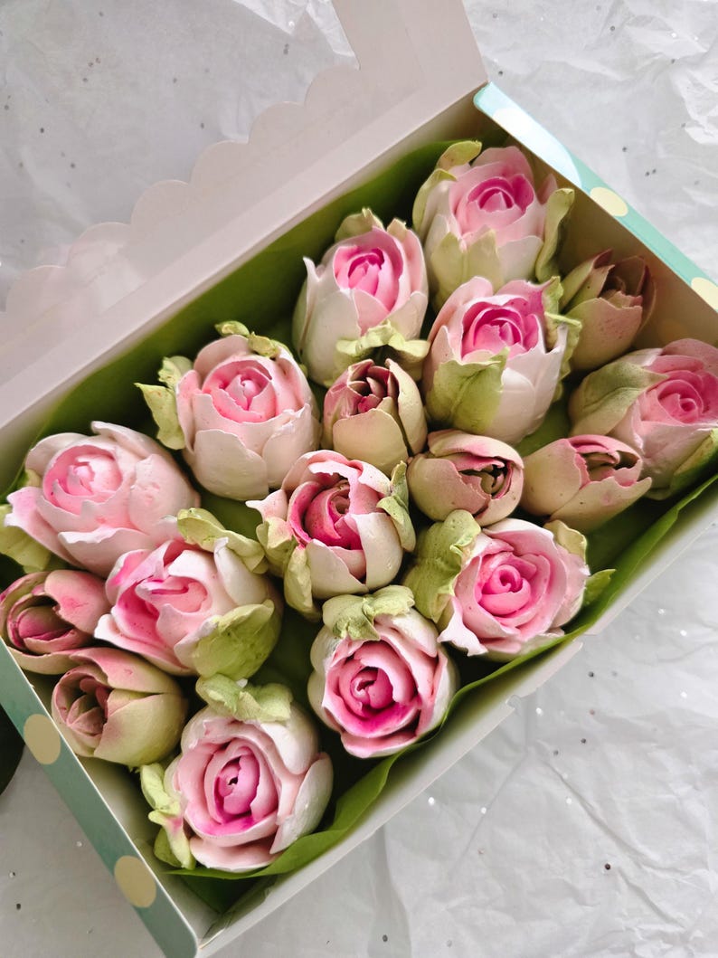 May include: A box of rose-shaped treats. The treats are pink and white, with green leaves. The box is white with a light green polka dot border. The treats are arranged on a green leaf-like paper.