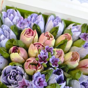May include: A box filled with edible flowers, featuring a variety of pastel-colored tulips and other blooms. The flowers are in shades of lavender, pink, and yellow, with green leaves. The arrangement is presented in a white box with a scalloped edge.