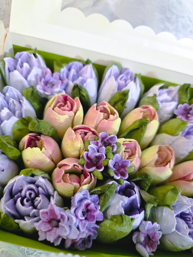 May include: A box filled with edible flowers, featuring a variety of pastel-colored tulips and other blooms. The flowers are in shades of lavender, pink, and yellow, with green leaves. The arrangement is presented in a white box with a scalloped edge.