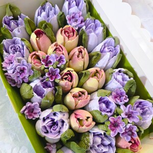 May include: A box filled with a colorful assortment of edible flowers. The arrangement features purple, pink, and yellow tulips and other floral elements with green leaves. The flowers are arranged in a white box with a scalloped edge.