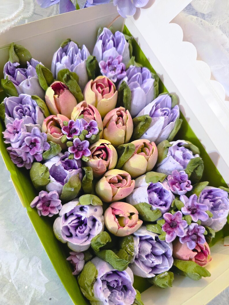 May include: A box filled with a colorful assortment of edible flowers. The arrangement features purple, pink, and yellow tulips and other floral elements with green leaves. The flowers are arranged in a white box with a scalloped edge.
