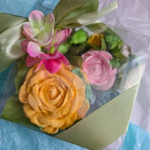 May include: A clear plastic box tied with a light green satin ribbon, containing a collection of colorful, handcrafted flowers. The flowers include a large yellow rose, pink roses, and other blossoms with green leaves.