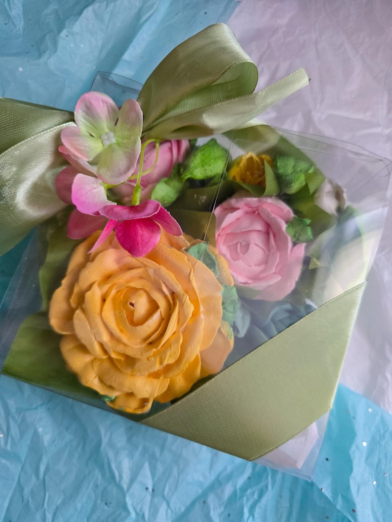 May include: A clear plastic box tied with a light green satin ribbon, containing a collection of colorful, handcrafted flowers. The flowers include a large yellow rose, pink roses, and other blossoms with green leaves.