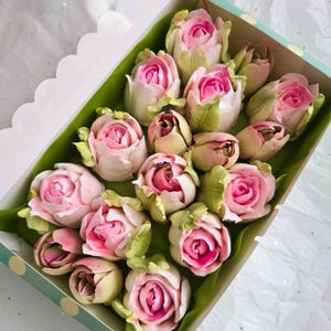 May include: A box of pink and white rose-shaped treats, possibly marshmallows or candies, arranged on green paper. The roses have a vibrant pink center and are nestled in a box with a polka dot design.