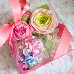 May include: A clear box tied with a pink ribbon, filled with colorful, handcrafted floral soaps. The soaps include a pink rose, a pink and blue hydrangea, and a rose with green and yellow hues. The box sits on a patterned paper.