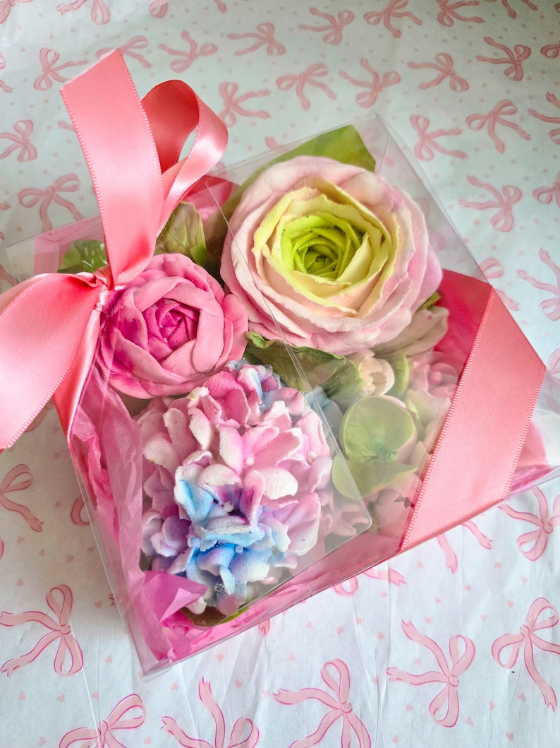 May include: A clear box tied with a pink ribbon, filled with colorful, handcrafted floral soaps. The soaps include a pink rose, a pink and blue hydrangea, and a rose with green and yellow hues. The box sits on a patterned paper.