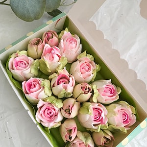 May include: A box of rose-shaped treats with pink and white petals and green leaves. The treats are arranged in a box with a clear window, and the box is decorated with a polka dot pattern.