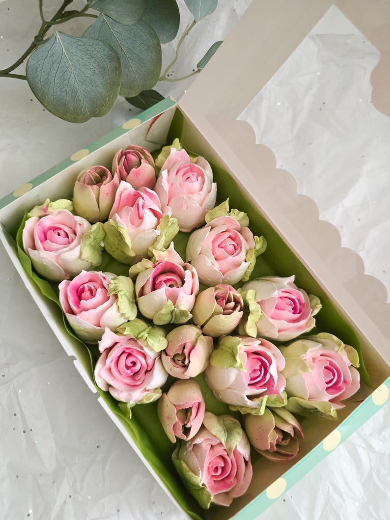 May include: A box of rose-shaped treats with pink and white petals and green leaves. The treats are arranged in a box with a clear window, and the box is decorated with a polka dot pattern.