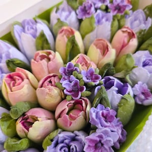 May include: A box filled with an assortment of colorful, handcrafted floral arrangements. The flowers feature shades of pink, purple, and white, with green leaves. The arrangement includes tulips and other flower types, creating a visually appealing display.