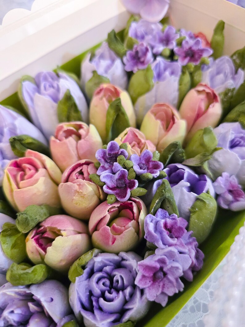 May include: A box filled with an assortment of colorful, handcrafted floral arrangements. The flowers feature shades of pink, purple, and white, with green leaves. The arrangement includes tulips and other flower types, creating a visually appealing display.